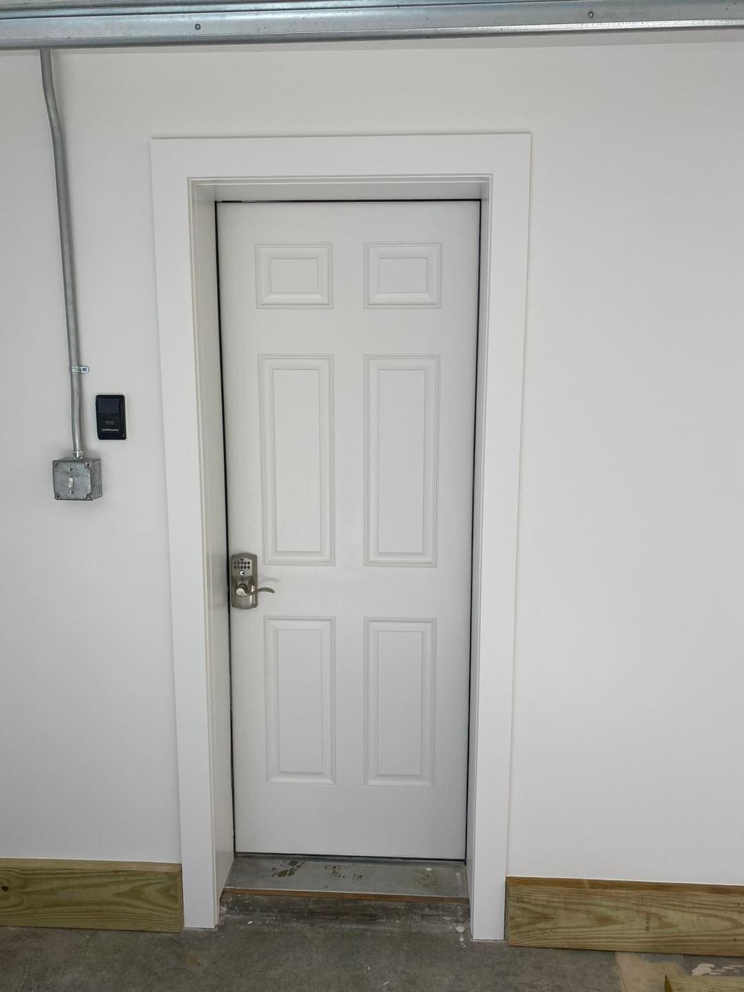 White door with gray hardware in a white-walled room with simple molding. Conduit and electrical box on the left.