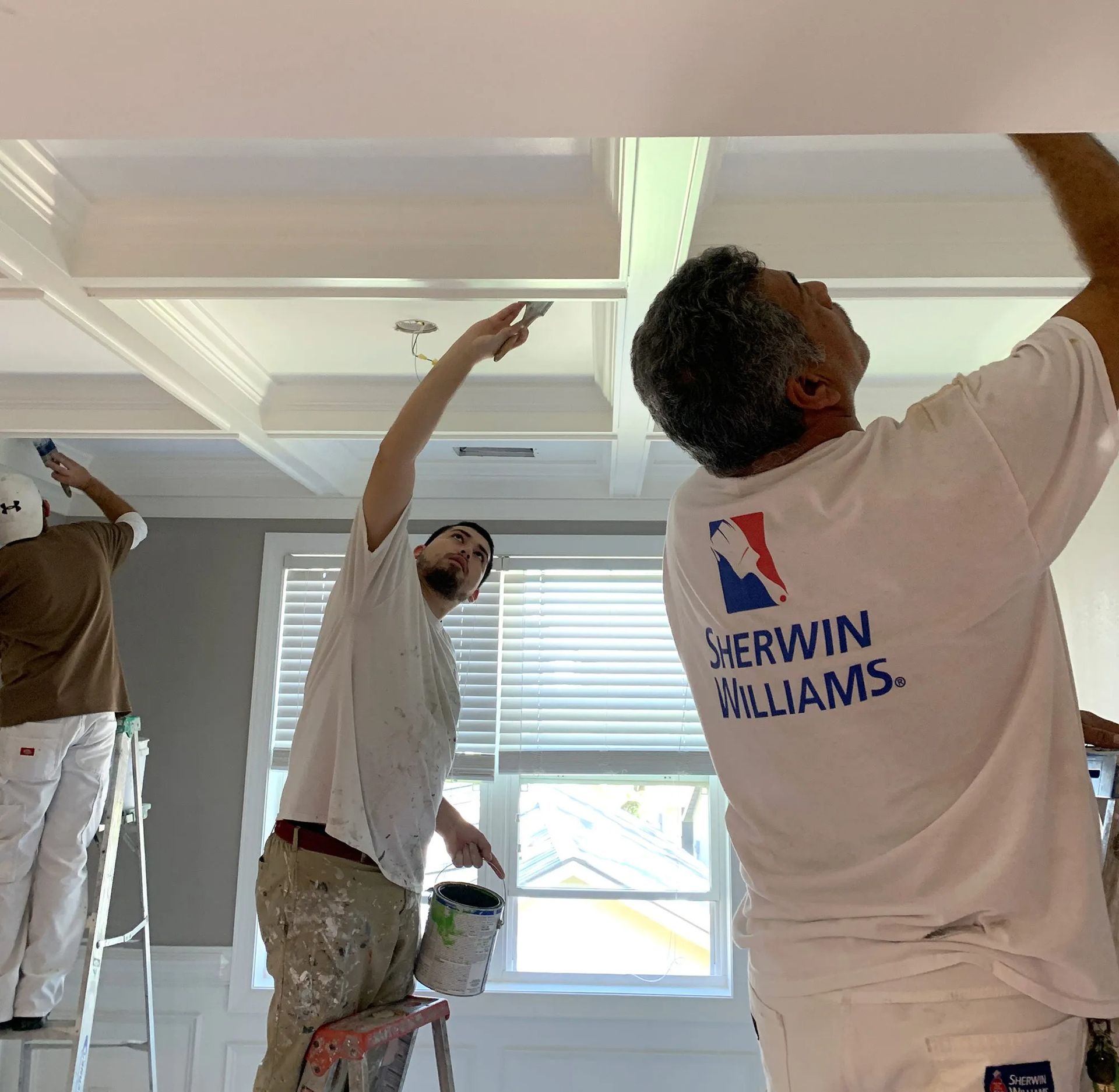 Three painters in white shirts painting a white ceiling in a bright room