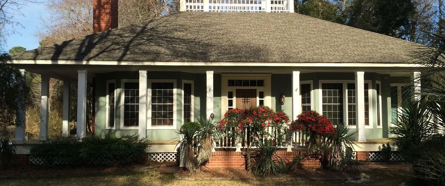 White house with a wide porch, dark roof, and flower boxes amid trees and shrubs.