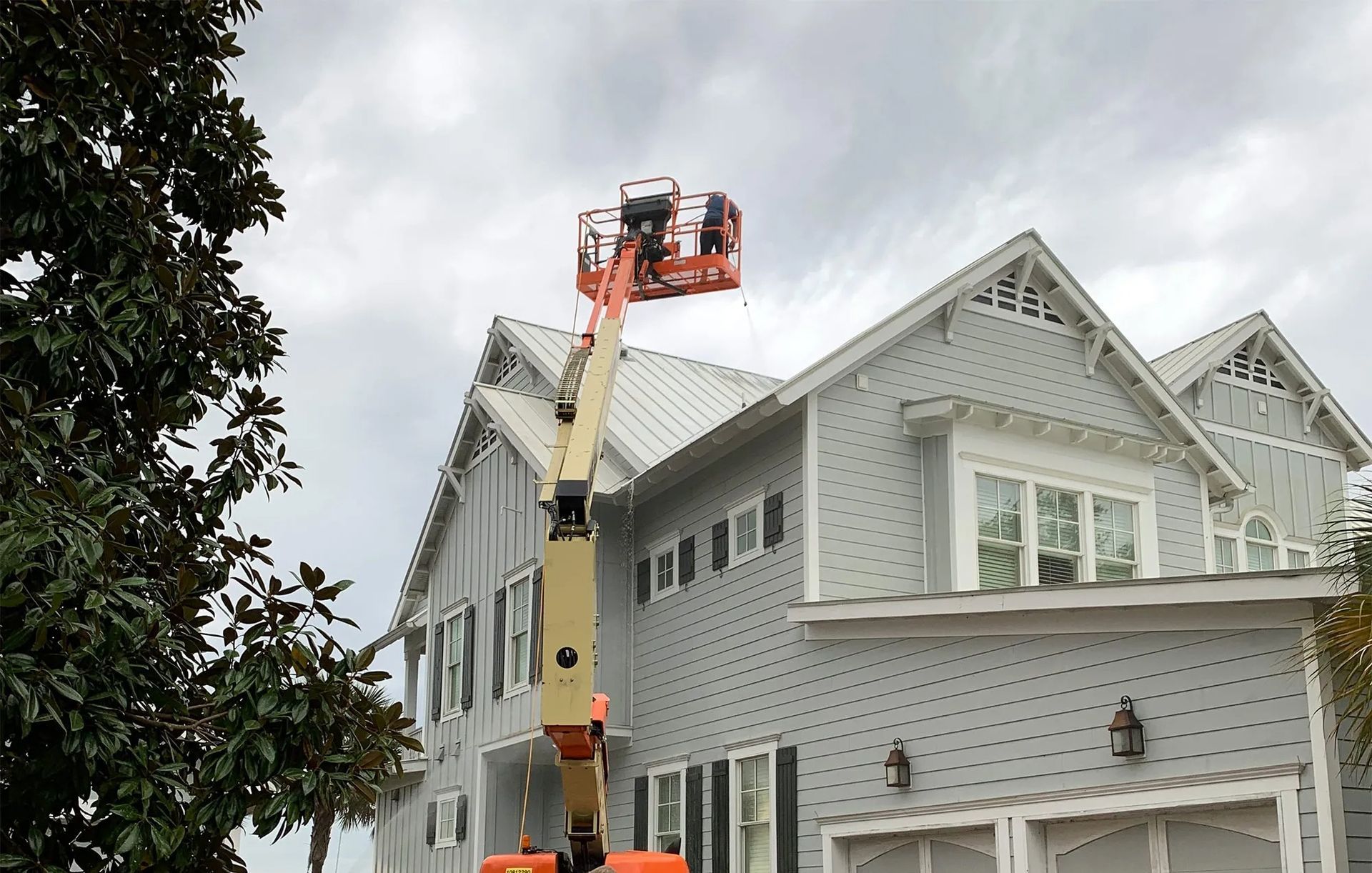 Worker in an orange lift inspecting a gray house’s roof and upper siding during repairs