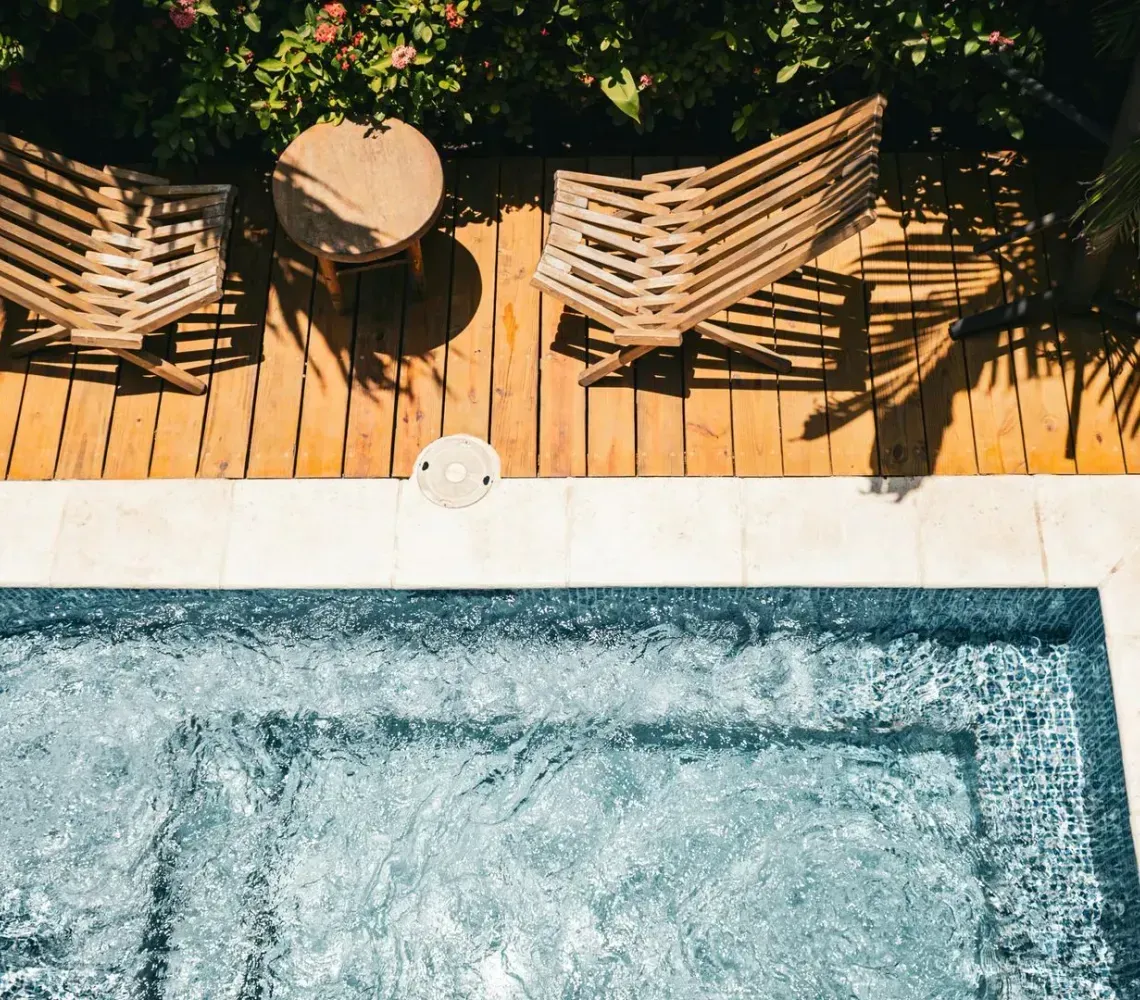 A small blue pool beside a wooden deck with two lounge chairs and tropical plants.
