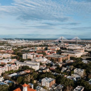 Aerial view of a city with dense buildings, roads, and a waterfront under a cloudy sky.
