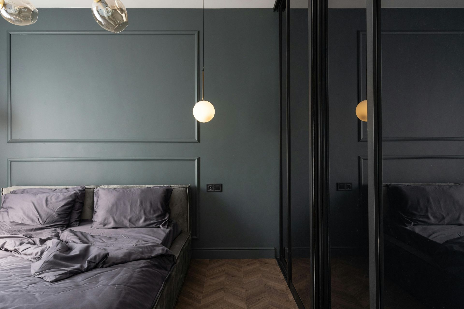 Modern gray bedroom with rumpled bed, paneled wall, hanging lights, and mirrored closet doors.