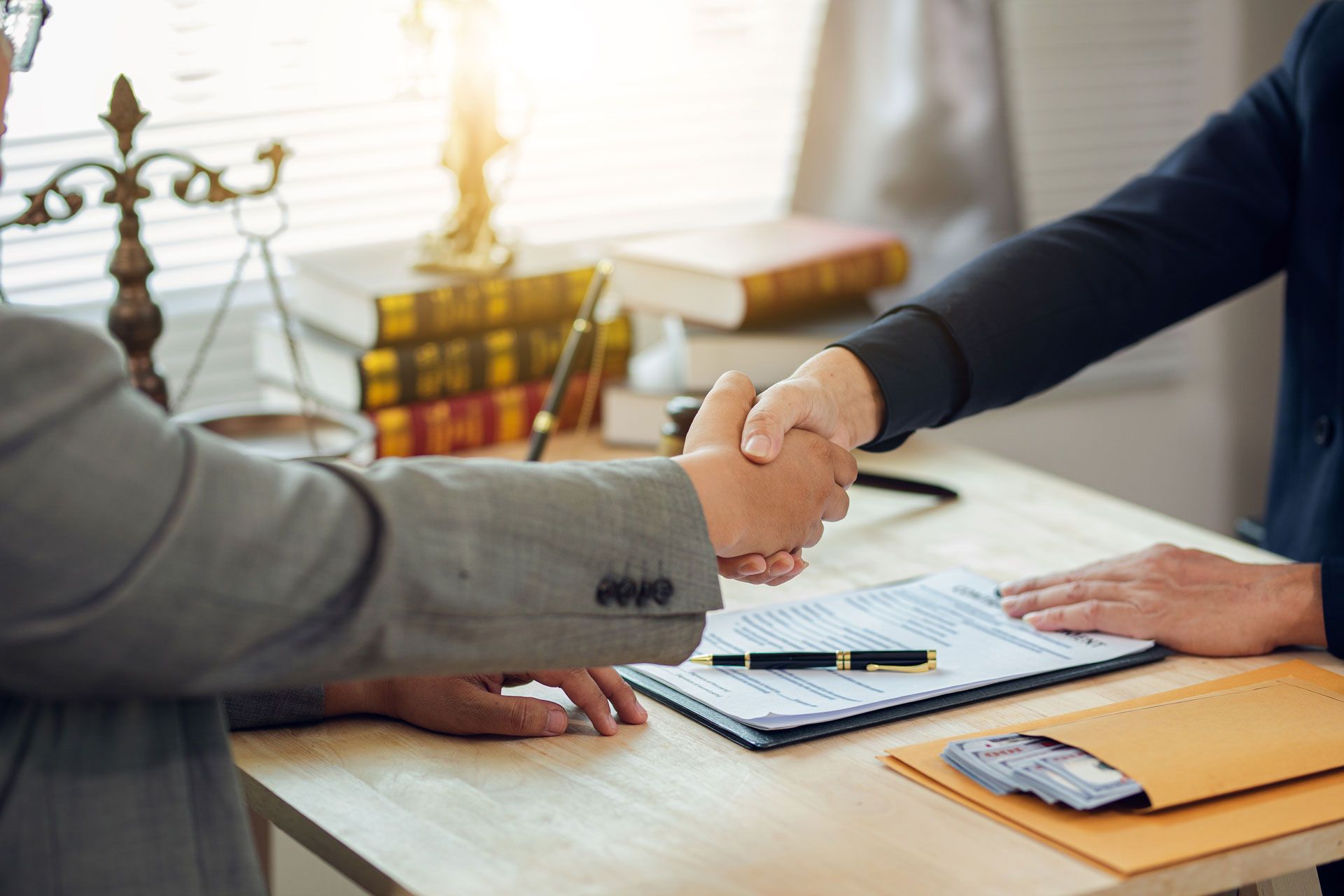 Two people shaking hands across a desk with paperwork, a calculator, and a laptop in a bright office.
