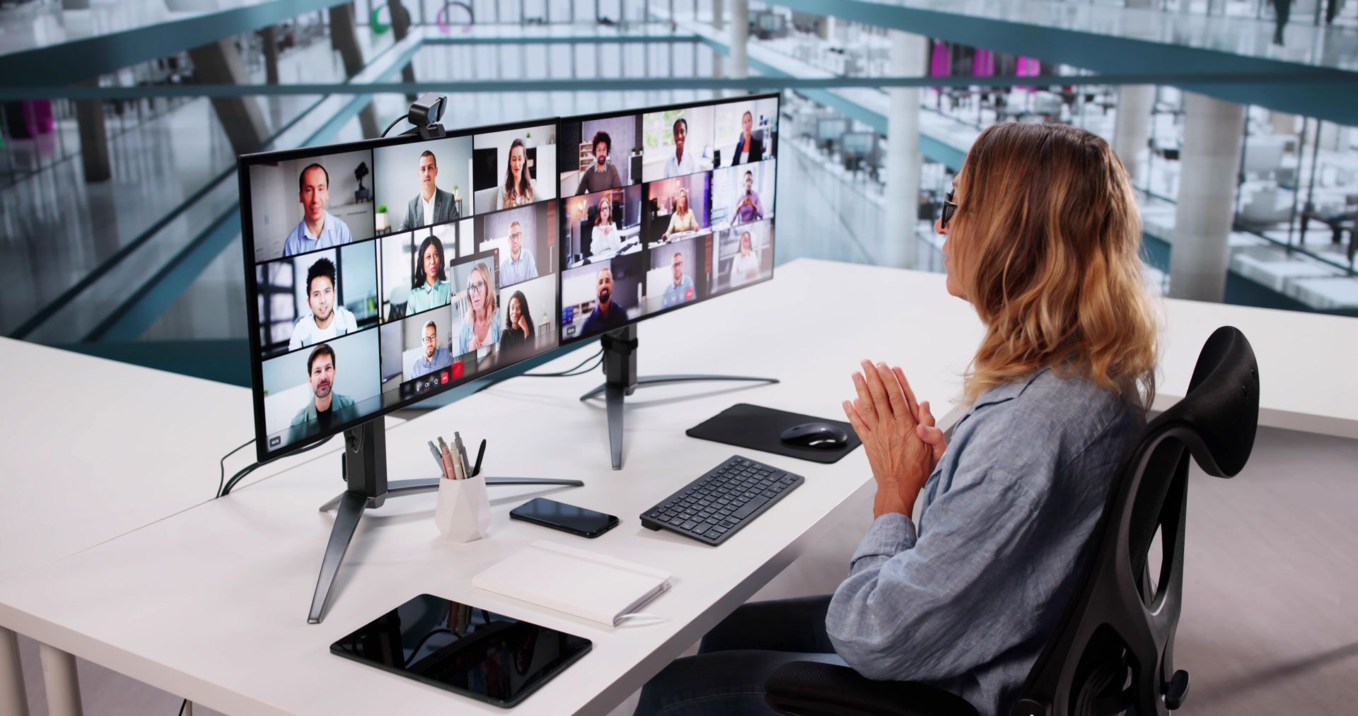 Person at a desk in a video meeting, facing multiple monitors with many participants visible.