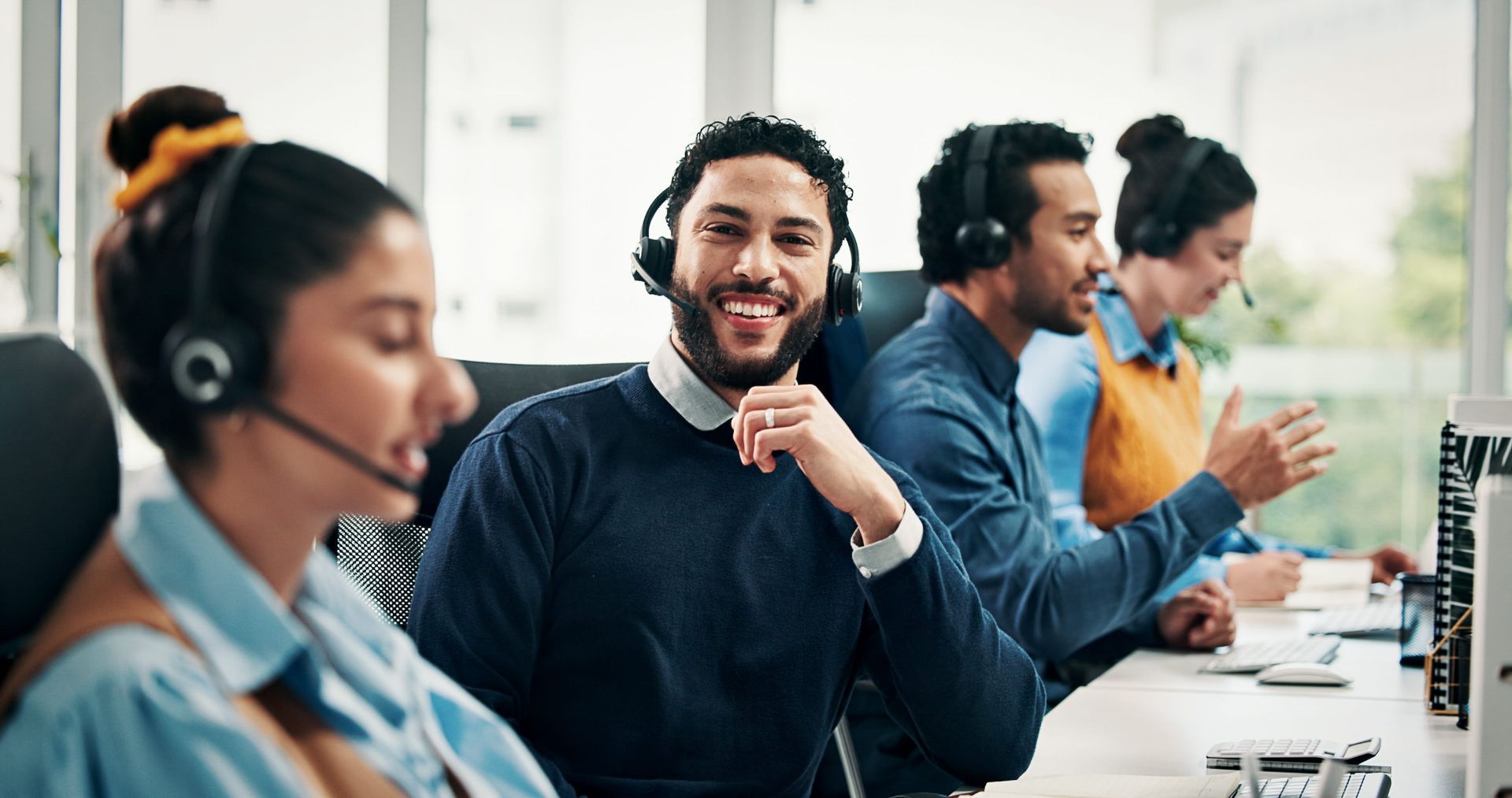 Call center agents wearing headsets in an office, one smiling at the camera while others work nearby