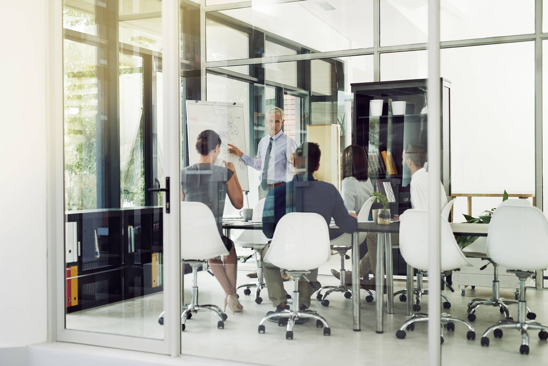 People in a bright glass conference room discussing around a table and white office chairs