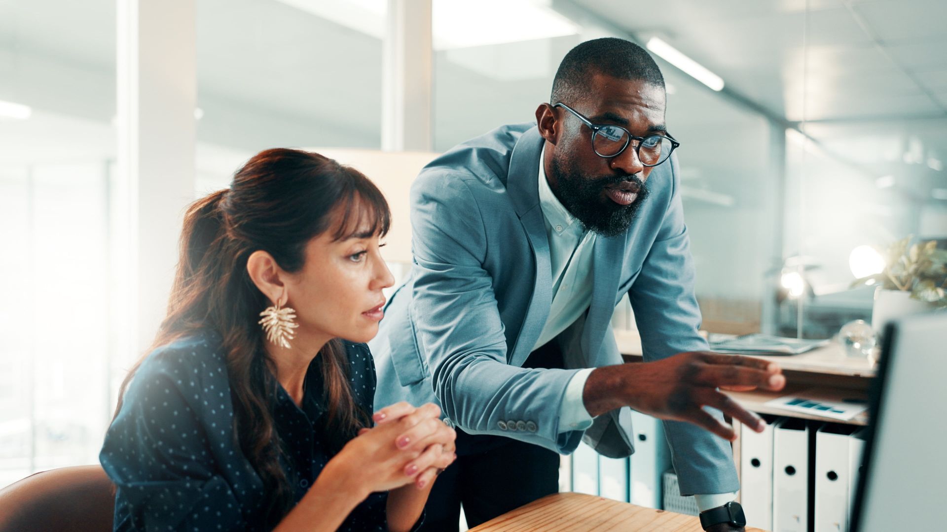 Two coworkers review a laptop in a bright office, one pointing at the screen.