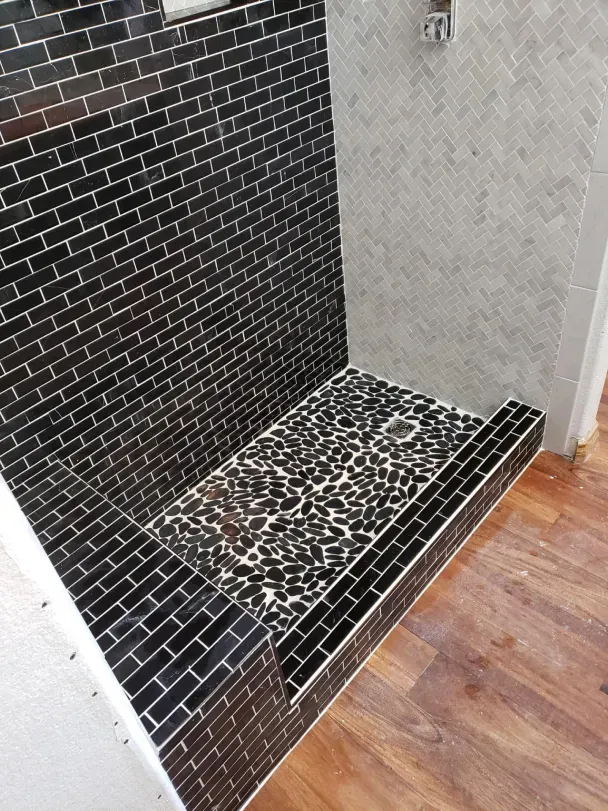 Black-tiled shower nook with pebble floor and light gray wall beside wood flooring