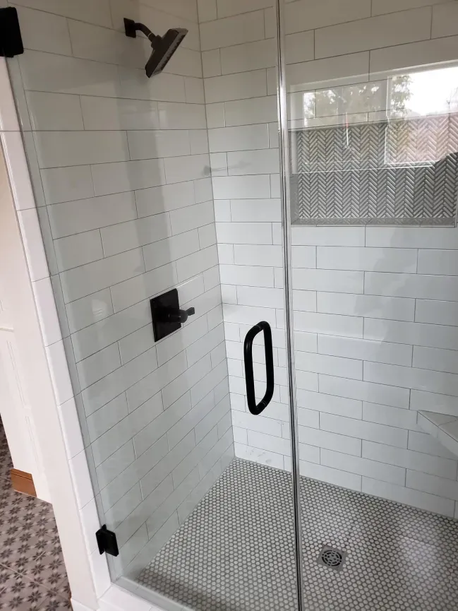 Glass-tiled shower with black fixtures, grab handle, and a bench under a window