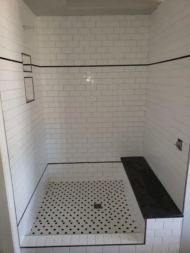 White-tiled walk-in shower with black accent trim, pebble floor, and built-in bench.
