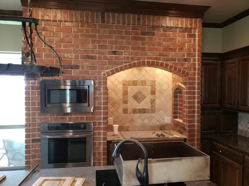 Kitchen with brick backsplash, built-in ovens, tiled alcove, and black sink in front of wooden cabinets