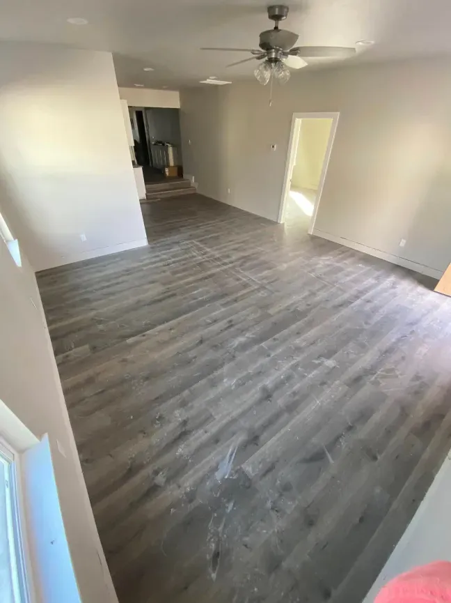 Empty living room with gray wood flooring, white walls, ceiling fan, and open doorway to another room