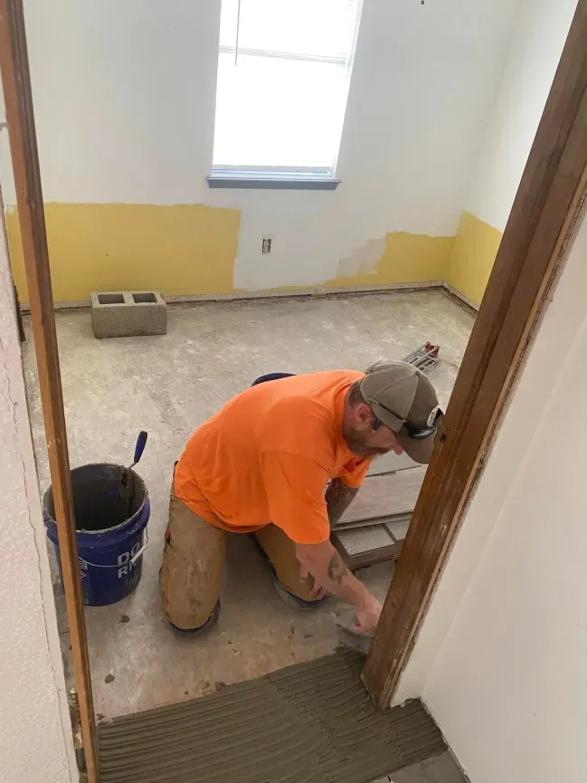 Worker troweling mortar on a floor in a small room with yellow walls and a window