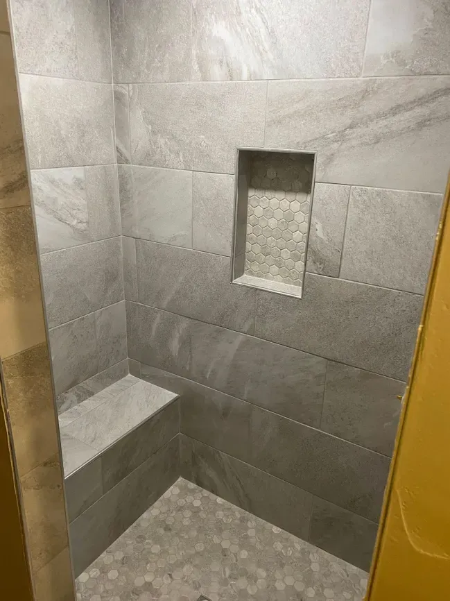 Small tiled shower nook with a built-in bench and recessed shelf in gray stone tile.