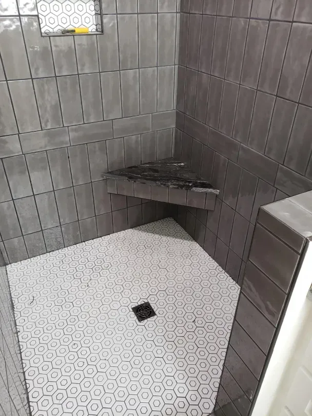 Tiled shower corner with a small built-in bench, hexagonal white floor tiles, and a center drain.