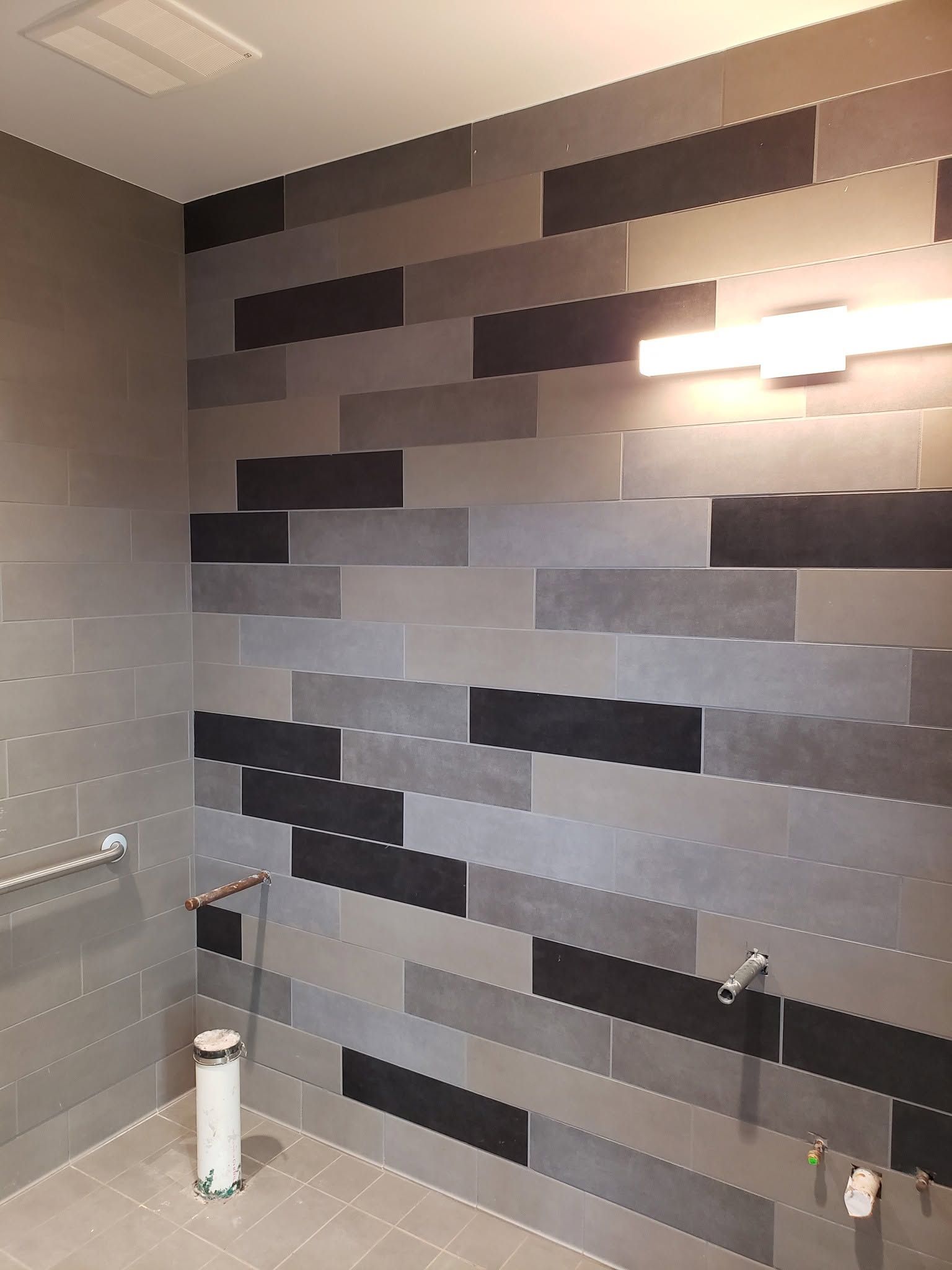 Gray tiled bathroom wall with a black-and-gray accent pattern and a bright wall light above a toilet paper holder