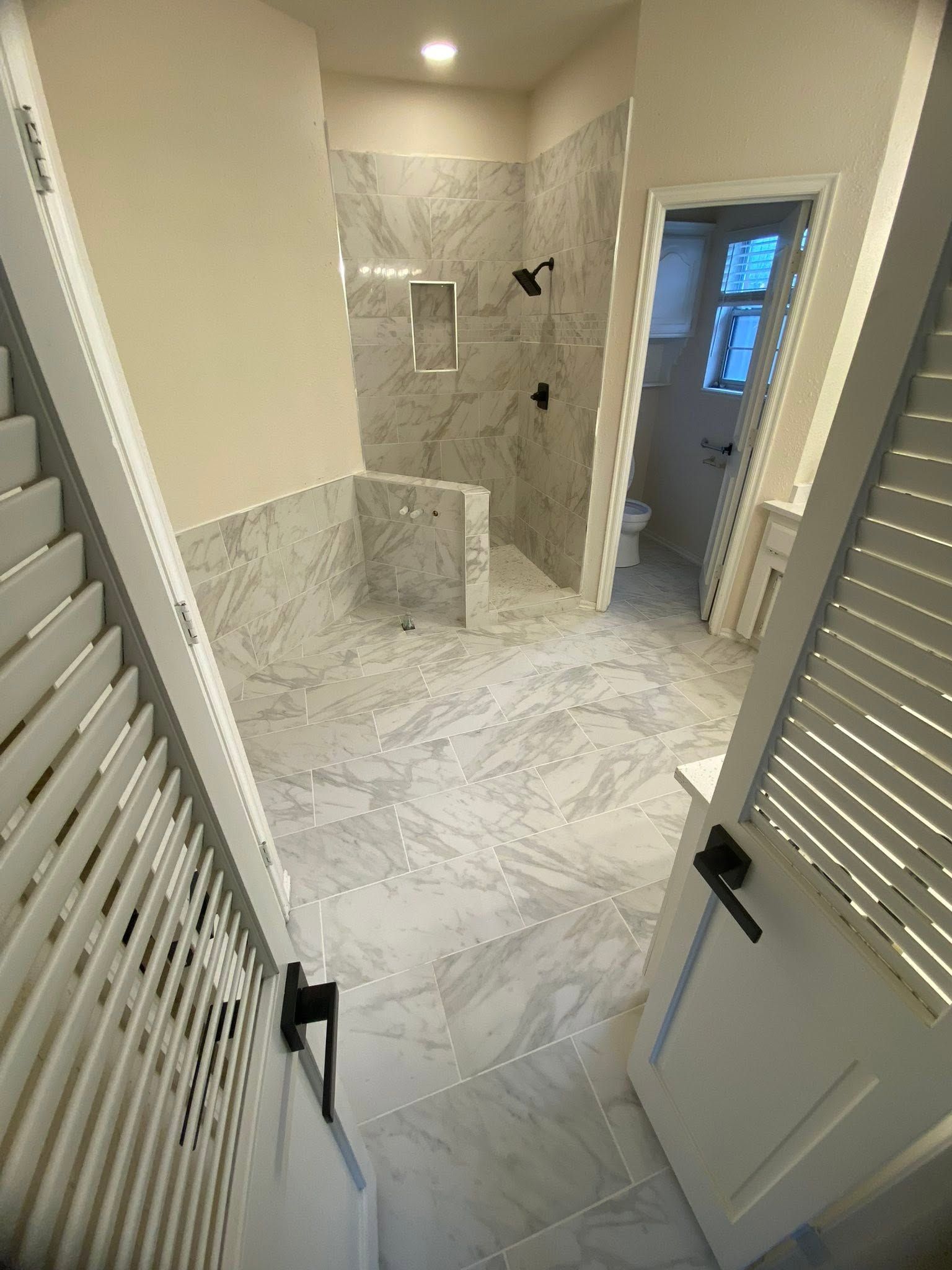 Bathroom with marble tile, walk-in shower, and open white doors in a bright hallway