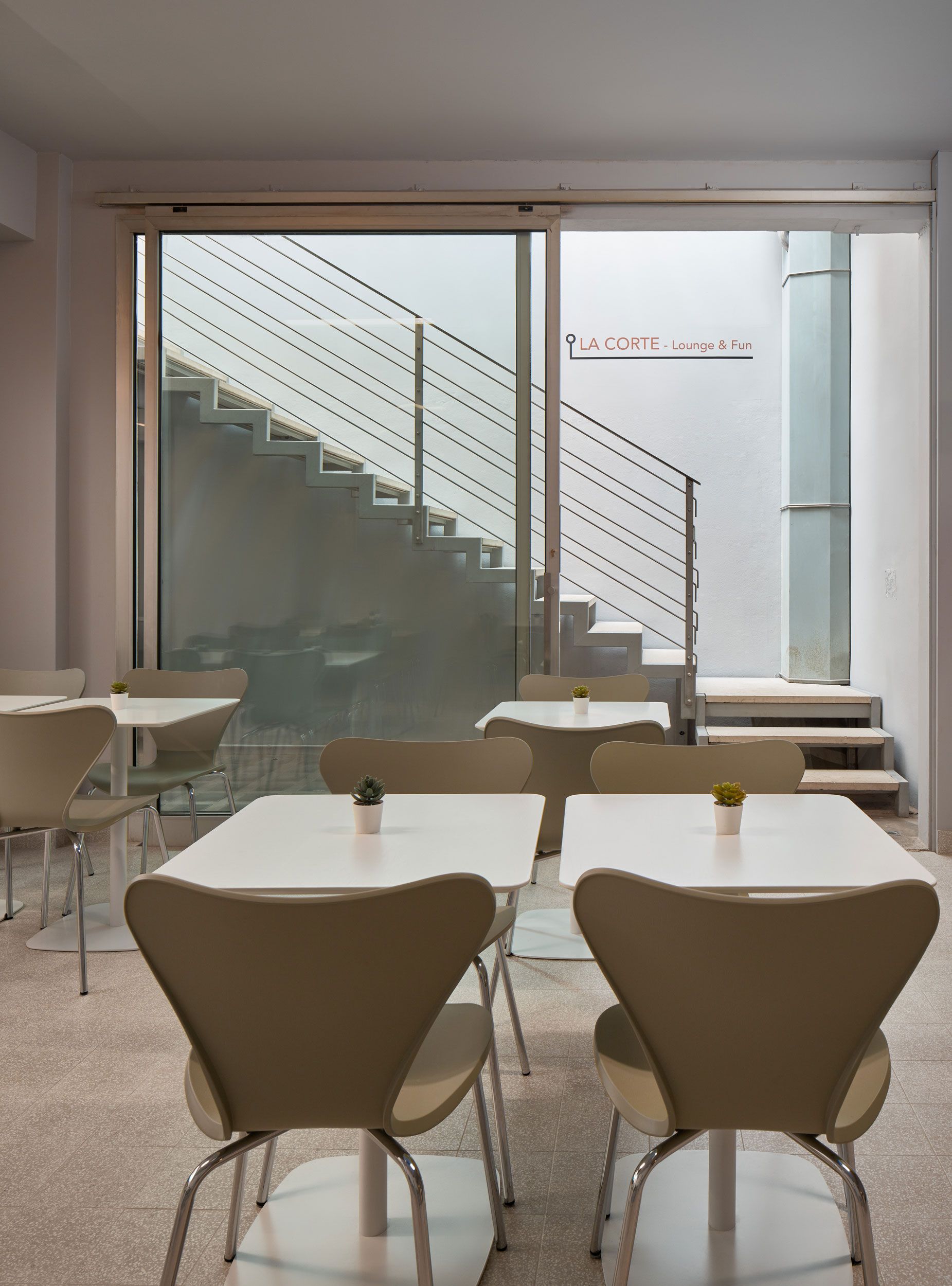 Interno di un bar con tavoli e sedie bianchi, una tromba delle scale visibile attraverso una porta a vetri.