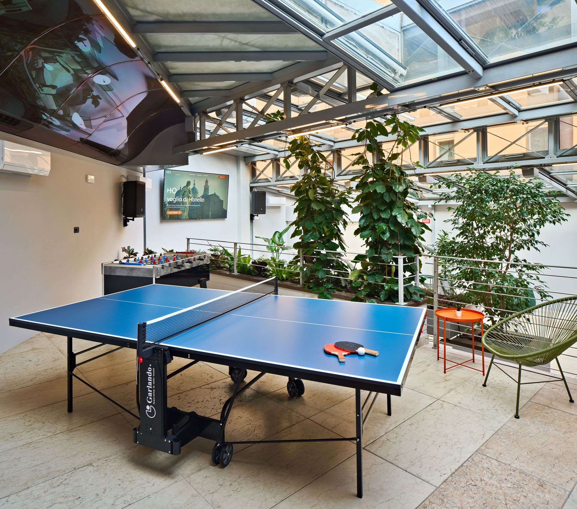 Tavolo da ping pong in una stanza soleggiata con biliardino, piante e posti a sedere.