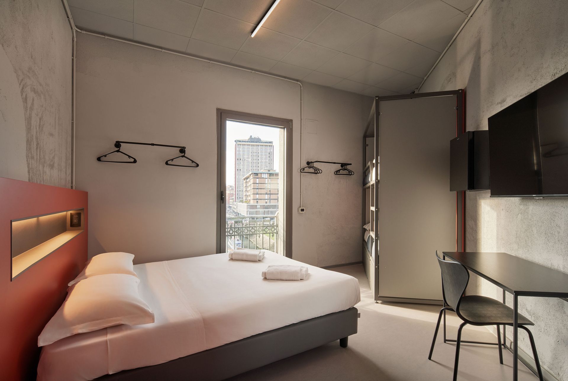 Moderna camera d'albergo con letto, scrivania, porta aperta con vista sulla città e due TV a parete.