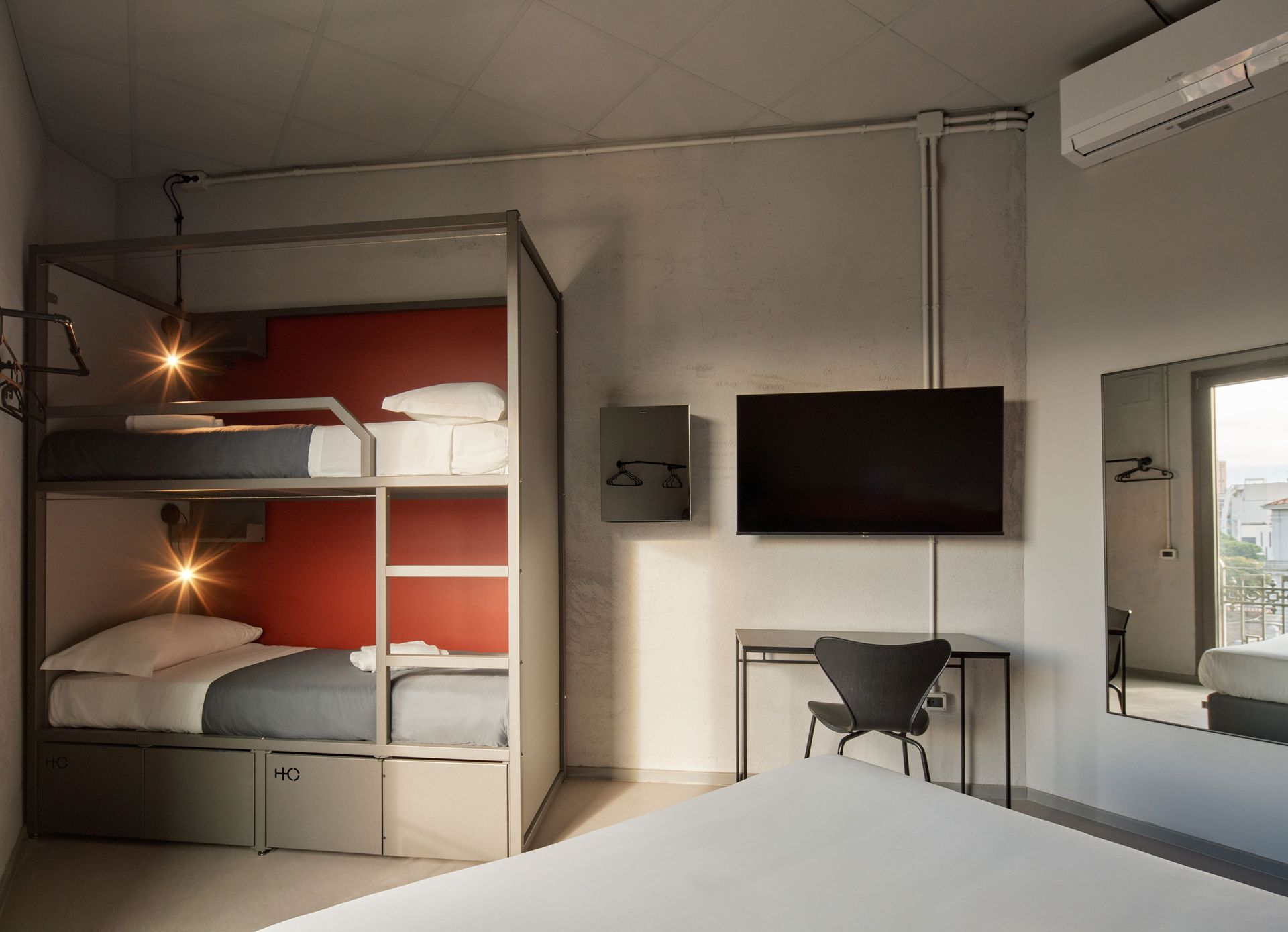 Moderna camera d'albergo con letti a castello, TV, scrivania e specchio. Colori bianco, grigio e rosso.