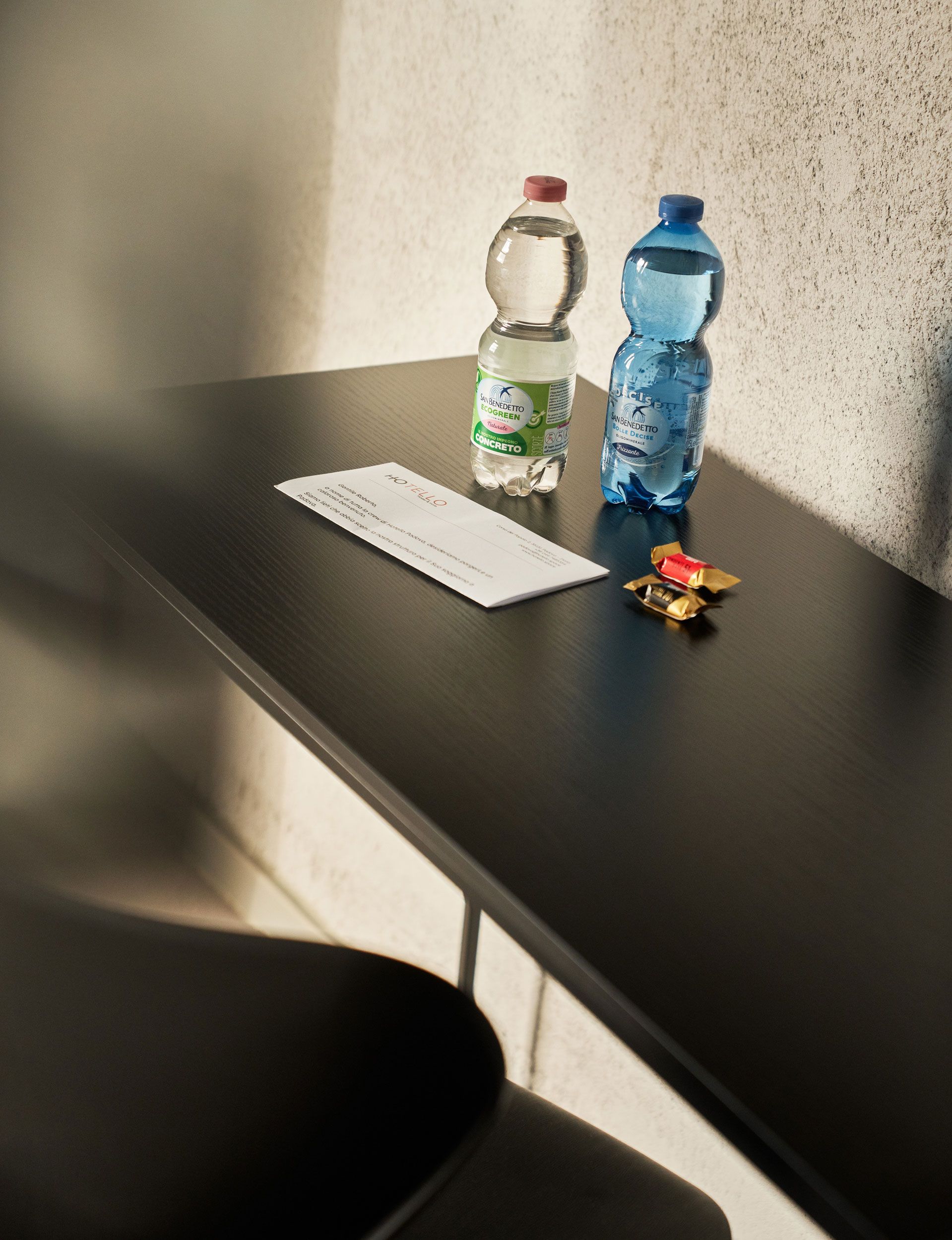 Tavolo buio con due bottiglie d'acqua, un foglio e delle chiavi. Luce del sole sul muro.