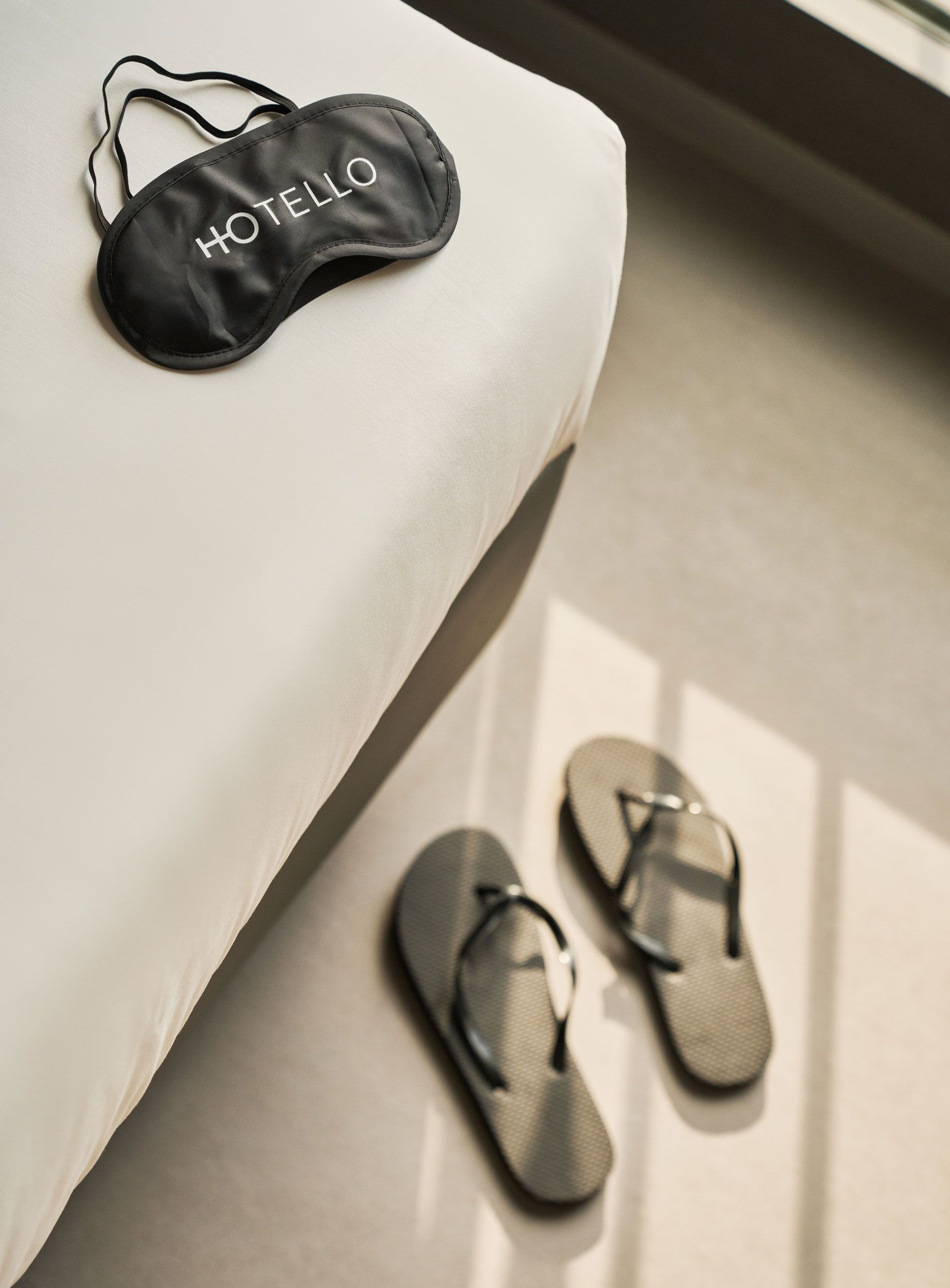 Mascherina per gli occhi e infradito su una superficie bianca, vicino a una finestra esposta alla luce del sole.