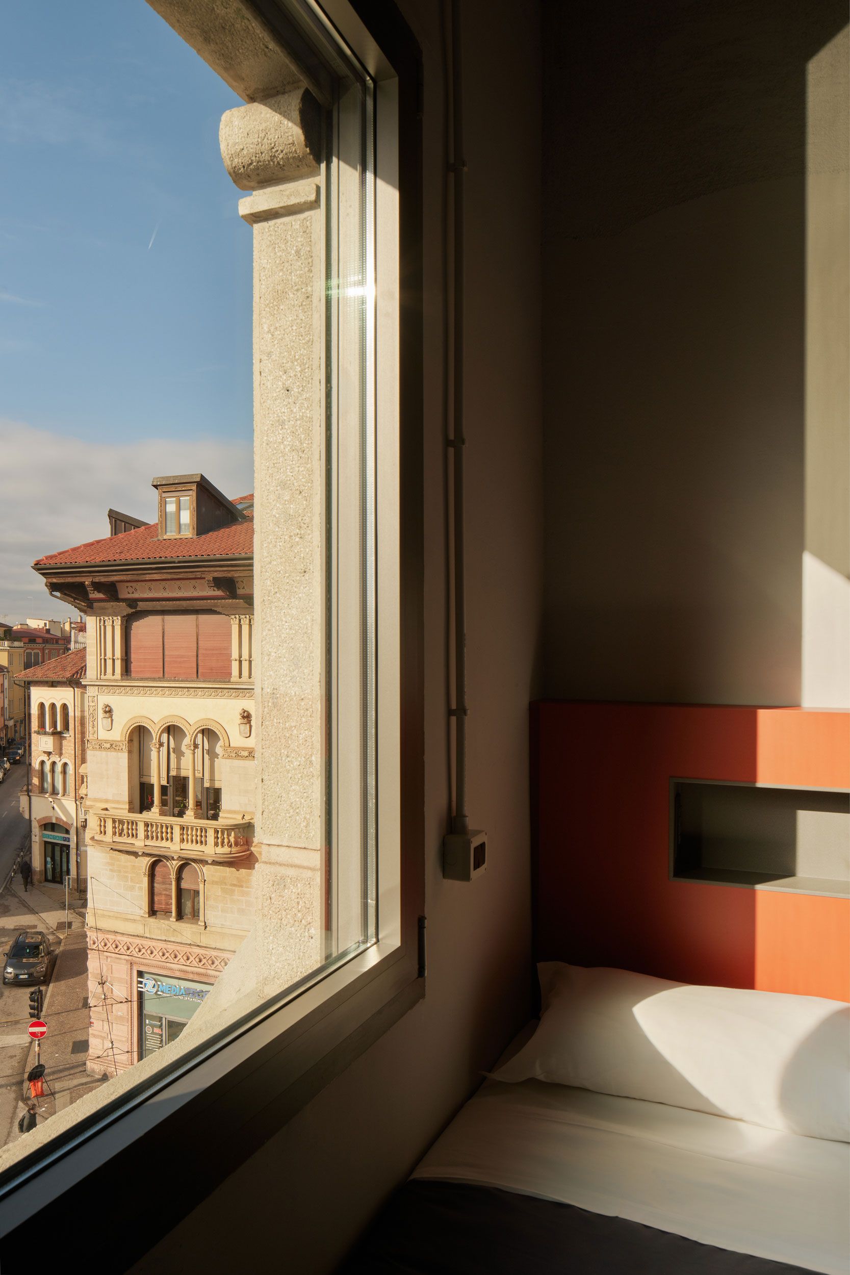 Camera soleggiata con letto e vista su una strada cittadina attraverso una finestra.