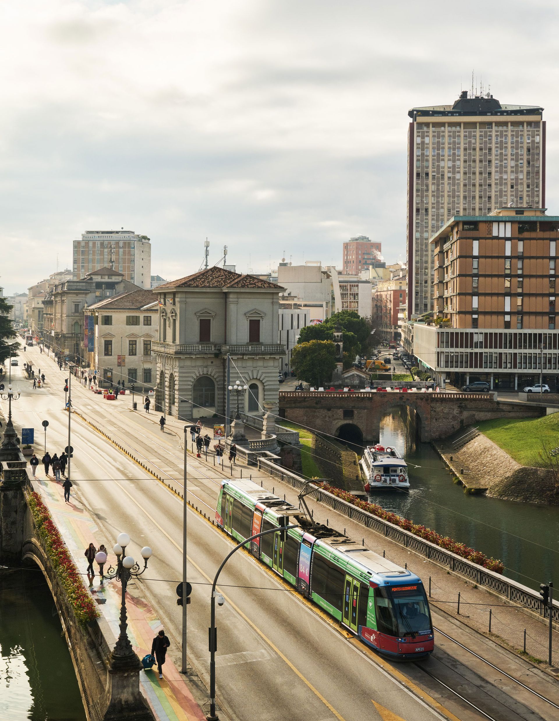 Paesaggio urbano con un tram su una strada larga, un ponte, acqua, edifici e cielo coperto.
