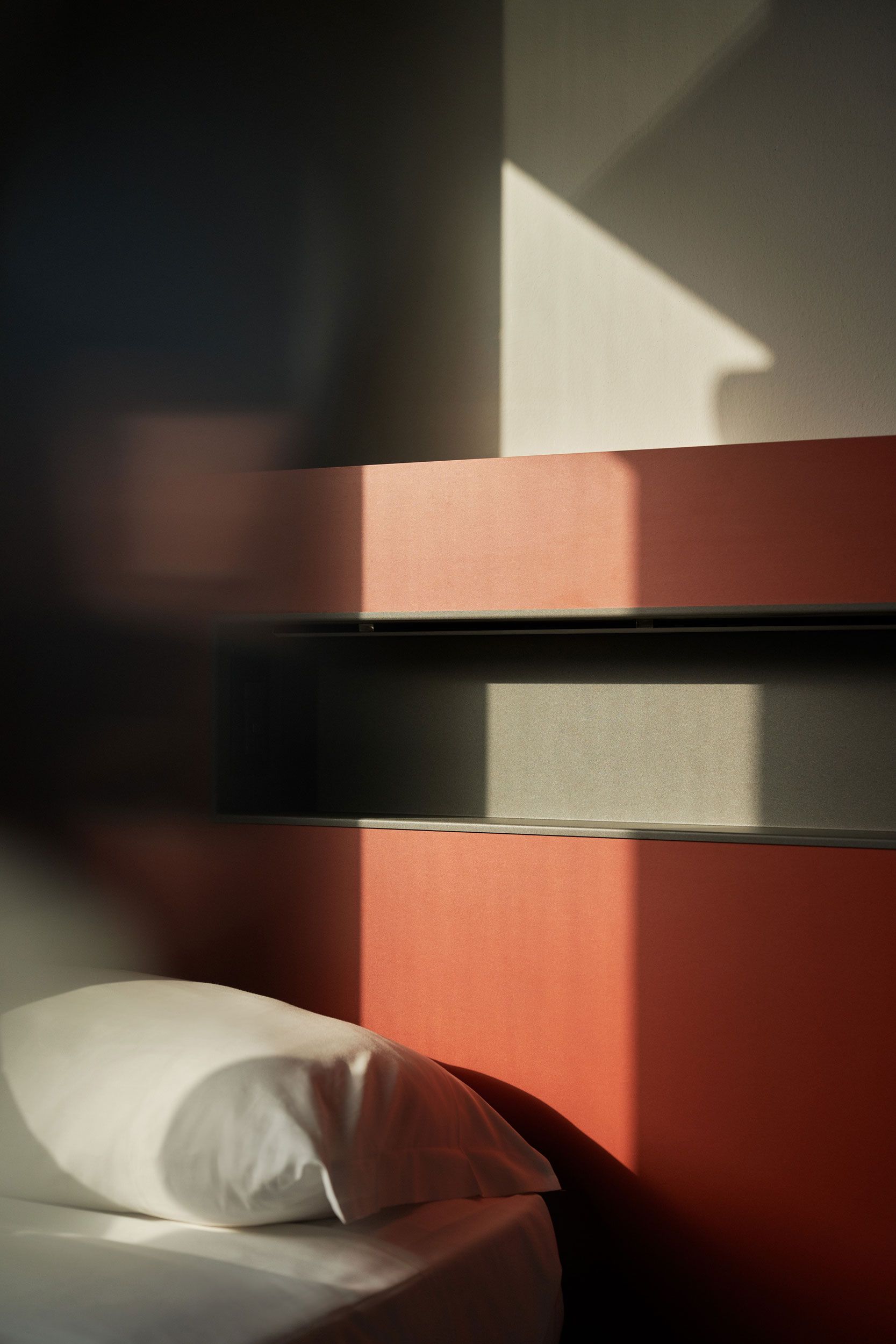 Letto con cuscino bianco di fronte a una parete geometrica arancione e grigia. La luce del sole crea ombre.
