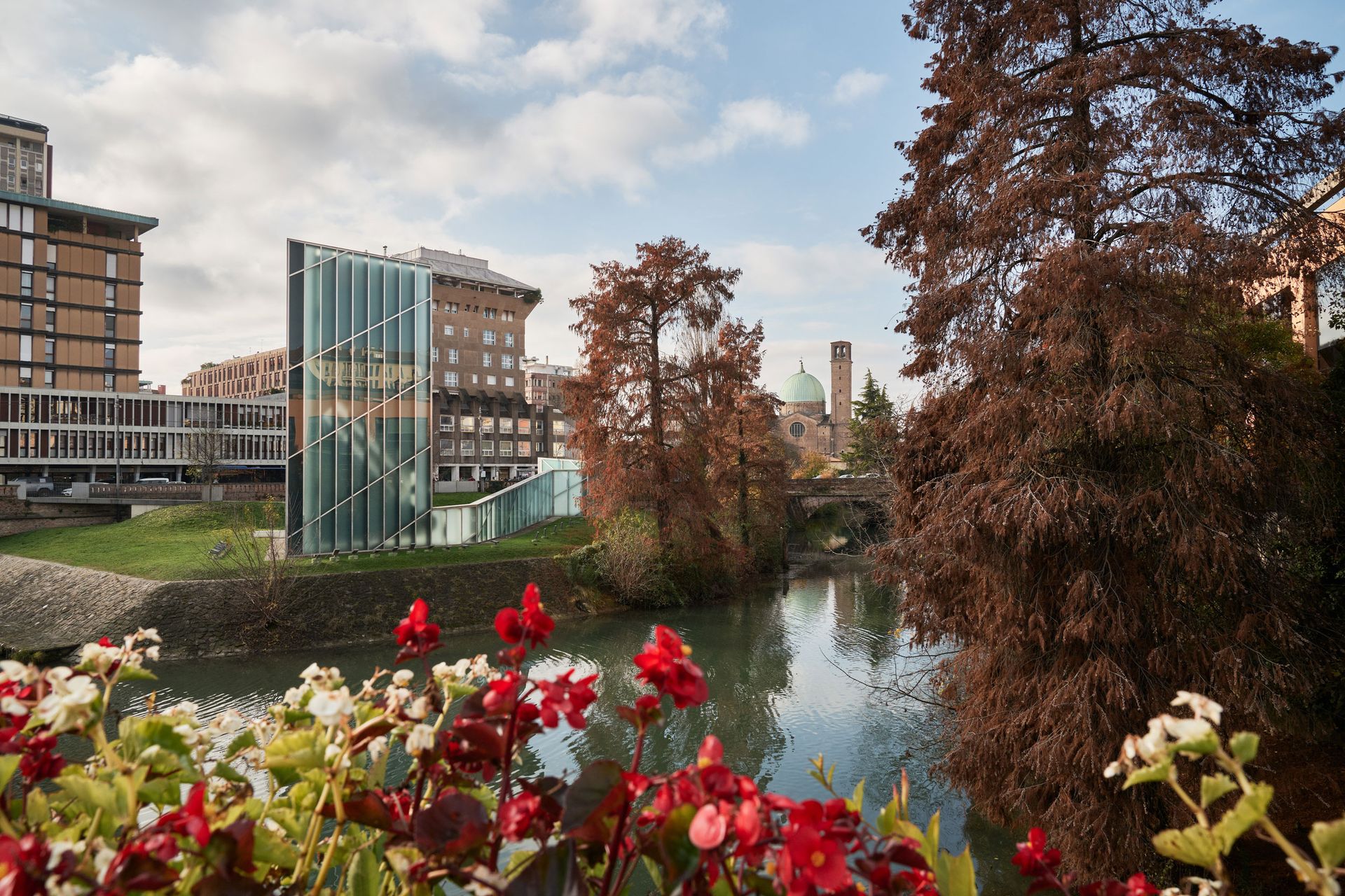 Un canale scorre tra edifici e alberi, con fiori rossi in primo piano. Cielo nuvoloso.