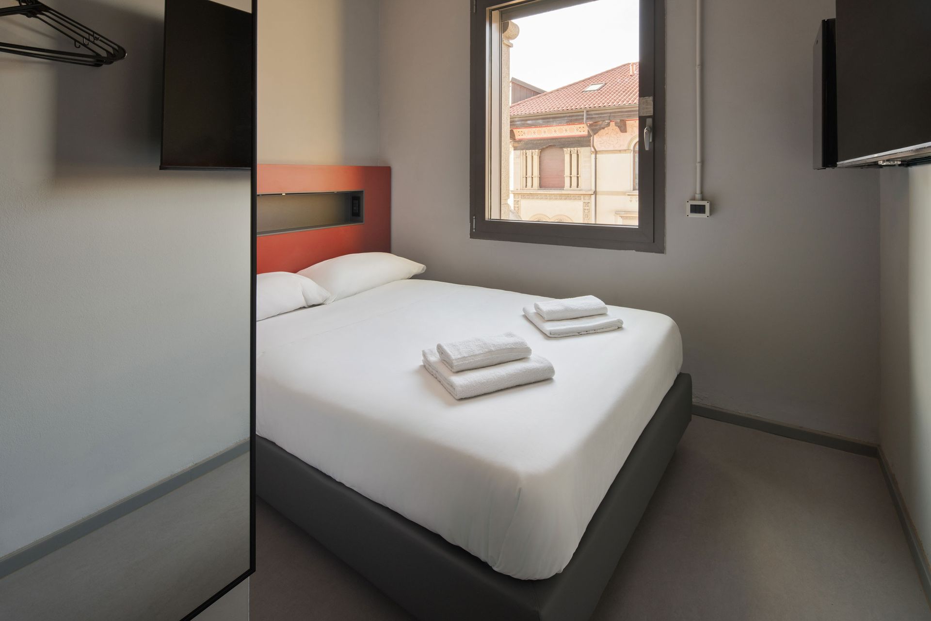 Piccola camera d'albergo con un letto, una finestra e una TV a parete.