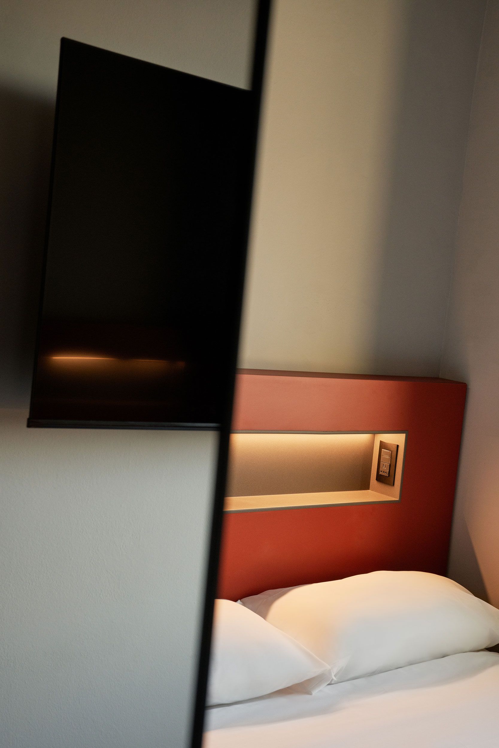 Una camera d'albergo con una TV montata a parete, una testiera rossa e un letto bianco.