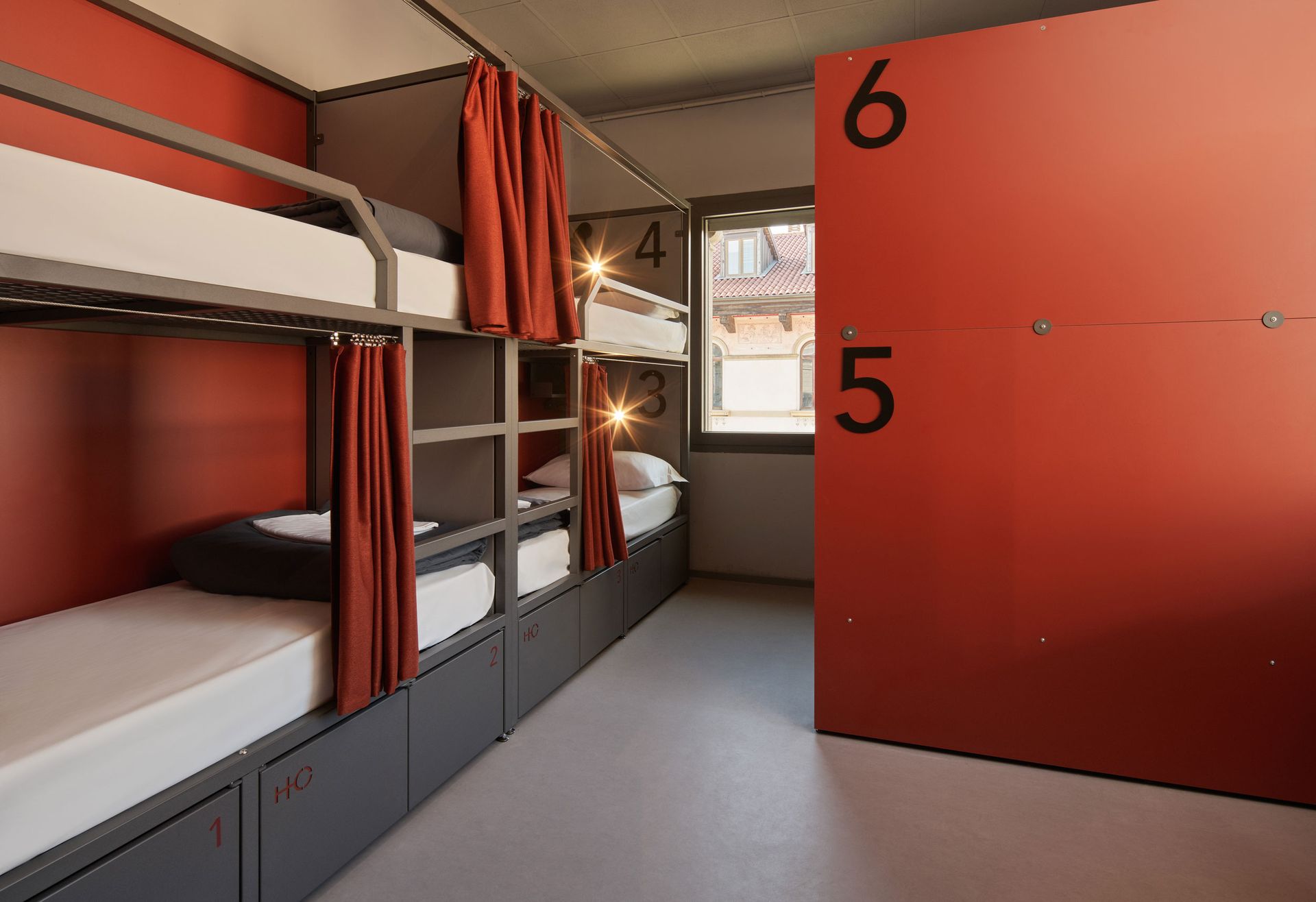 Stanza con letti a castello, letti rossi e grigi e armadietti. I numeri 5 e 6 sono visibili su un armadietto.