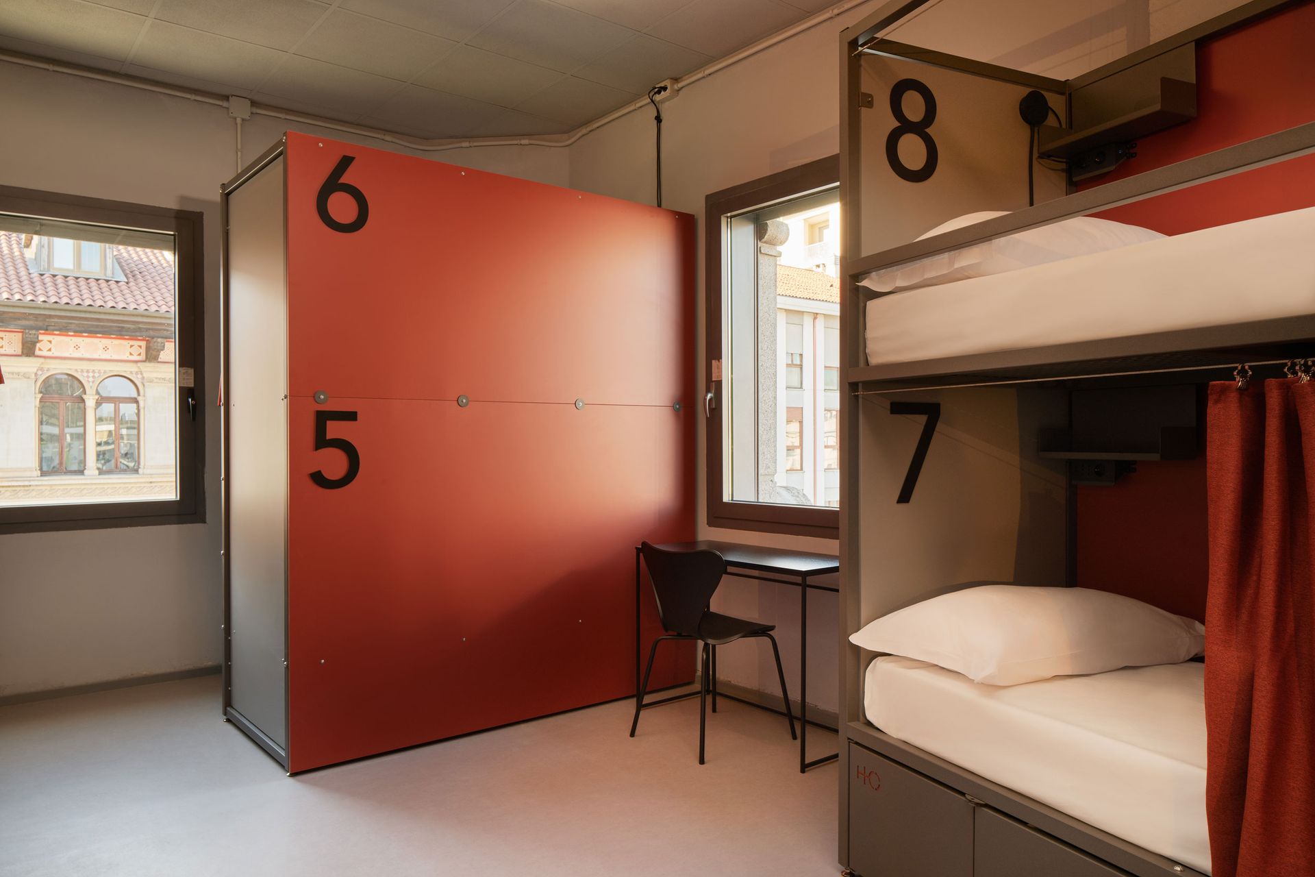 Camera in ostello con letti a castello e cabine numerate. Toni del rosso e del grigio, vista dalla finestra.