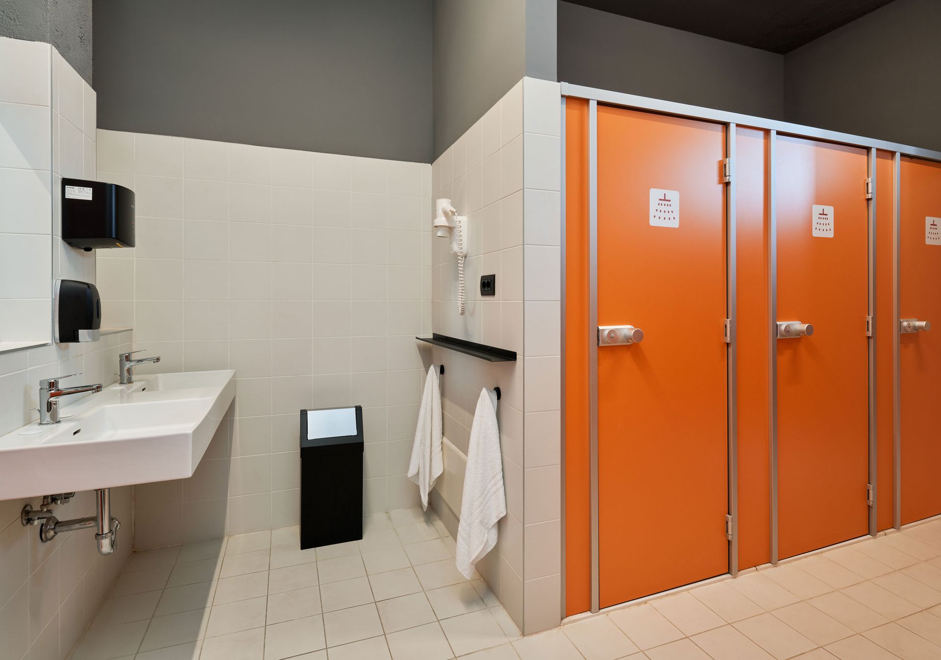 Bagno pubblico con porte arancioni, lavandini bianchi e pareti grigie.