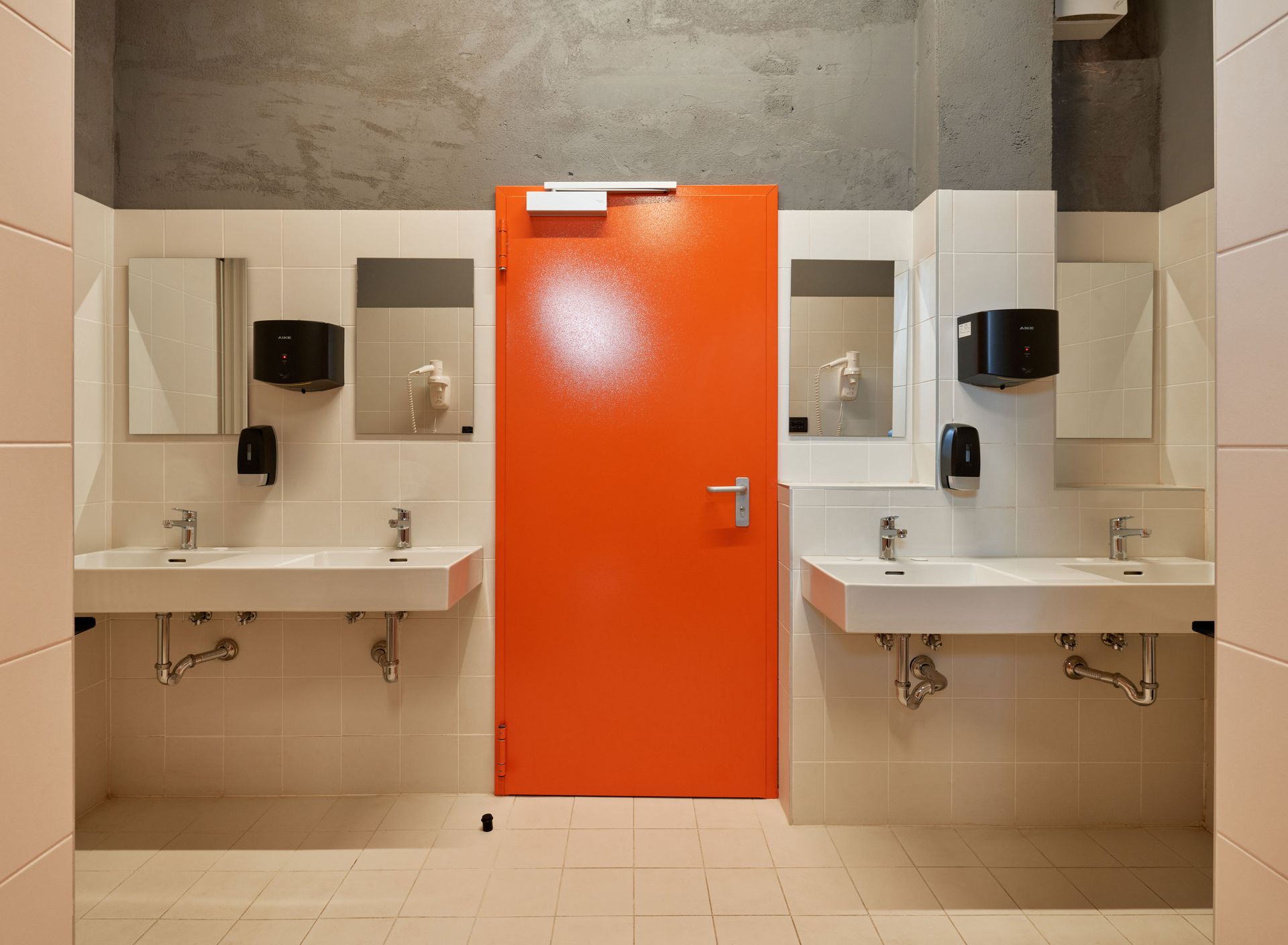 Bagno pubblico con lavandini bianchi, specchi e porta arancione.