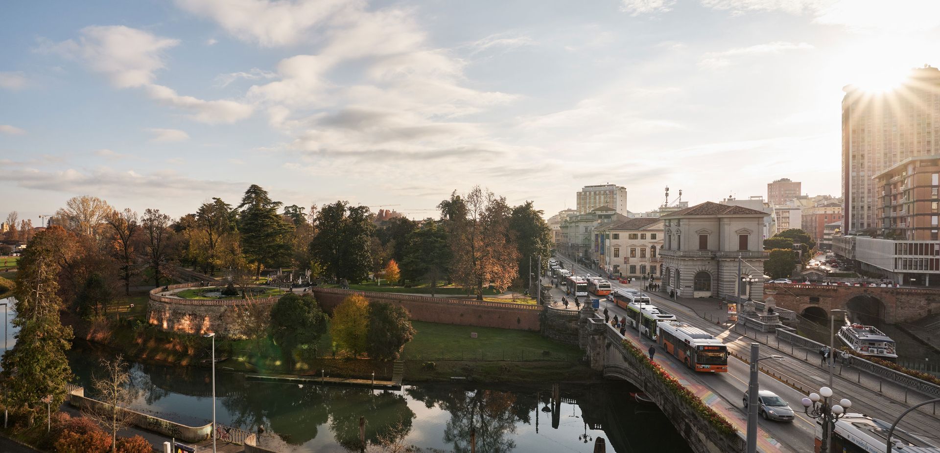 Un paesaggio urbano con un ponte su un fiume, un tram, edifici e un parco sotto un cielo nuvoloso.