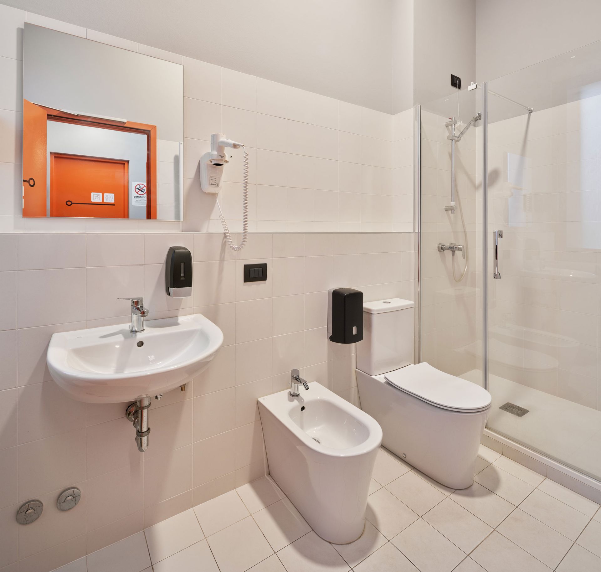 Bagno bianco con lavandino, bidet, WC, doccia e uno specchio con il riflesso arancione della porta.