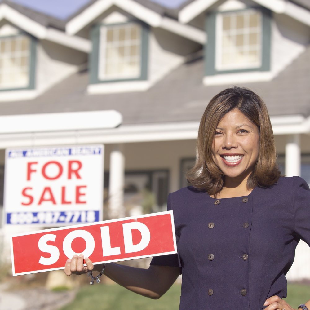 A woman holding a sold sign in front of a for sale sign