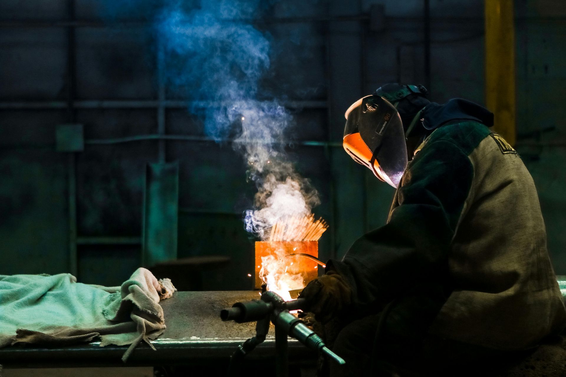 Welder in protective gear at work, creating sparks and smoke in a dark industrial setting.