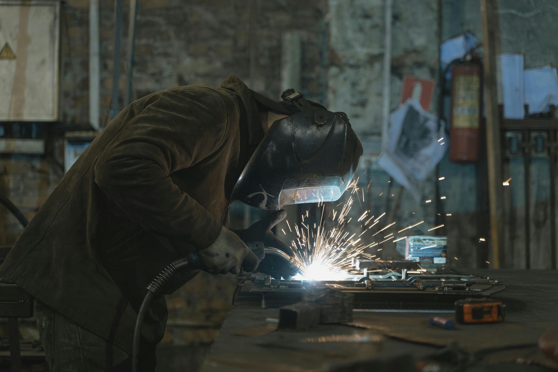 Welder wearing a mask and gloves, welding metal with bright sparks in a workshop.