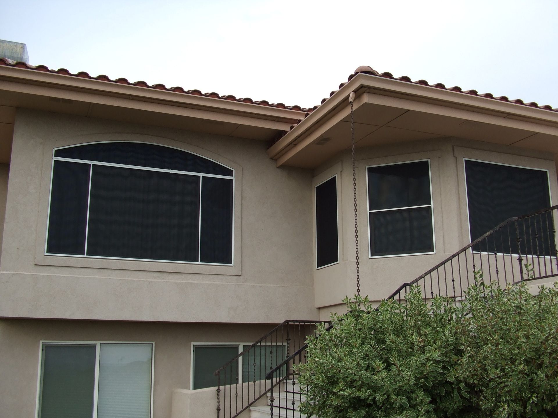 Two-story beige stucco house with large dark windows and a sloped roof behind bushes