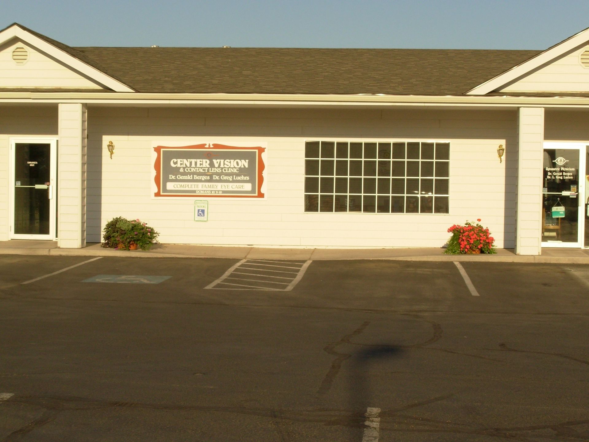 Single-story white storefront with large window, sign, and small flower beds beside an empty parking lot