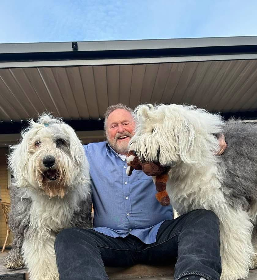 Man sitting between two shaggy white dogs under a patio roof, all facing the camera.