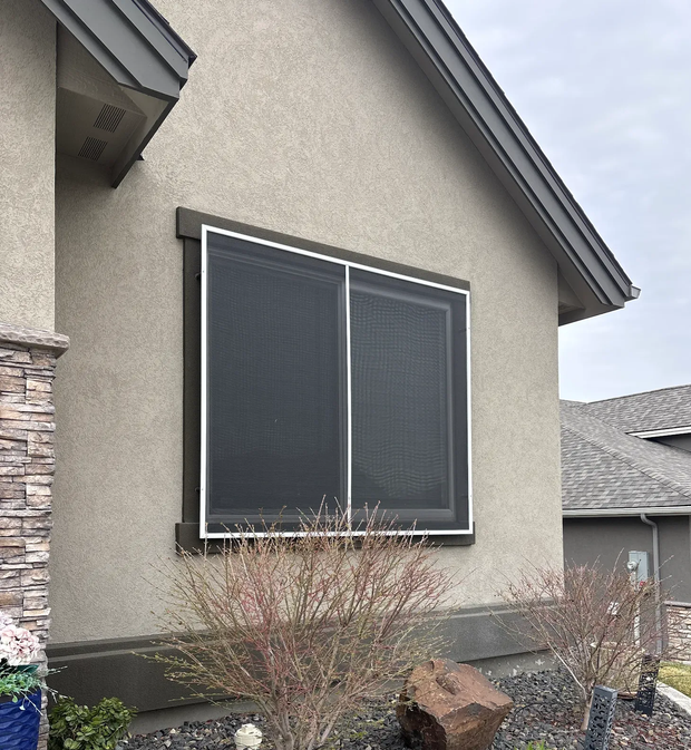Exterior of a stucco house with a large dark window and landscaped shrubs beneath it