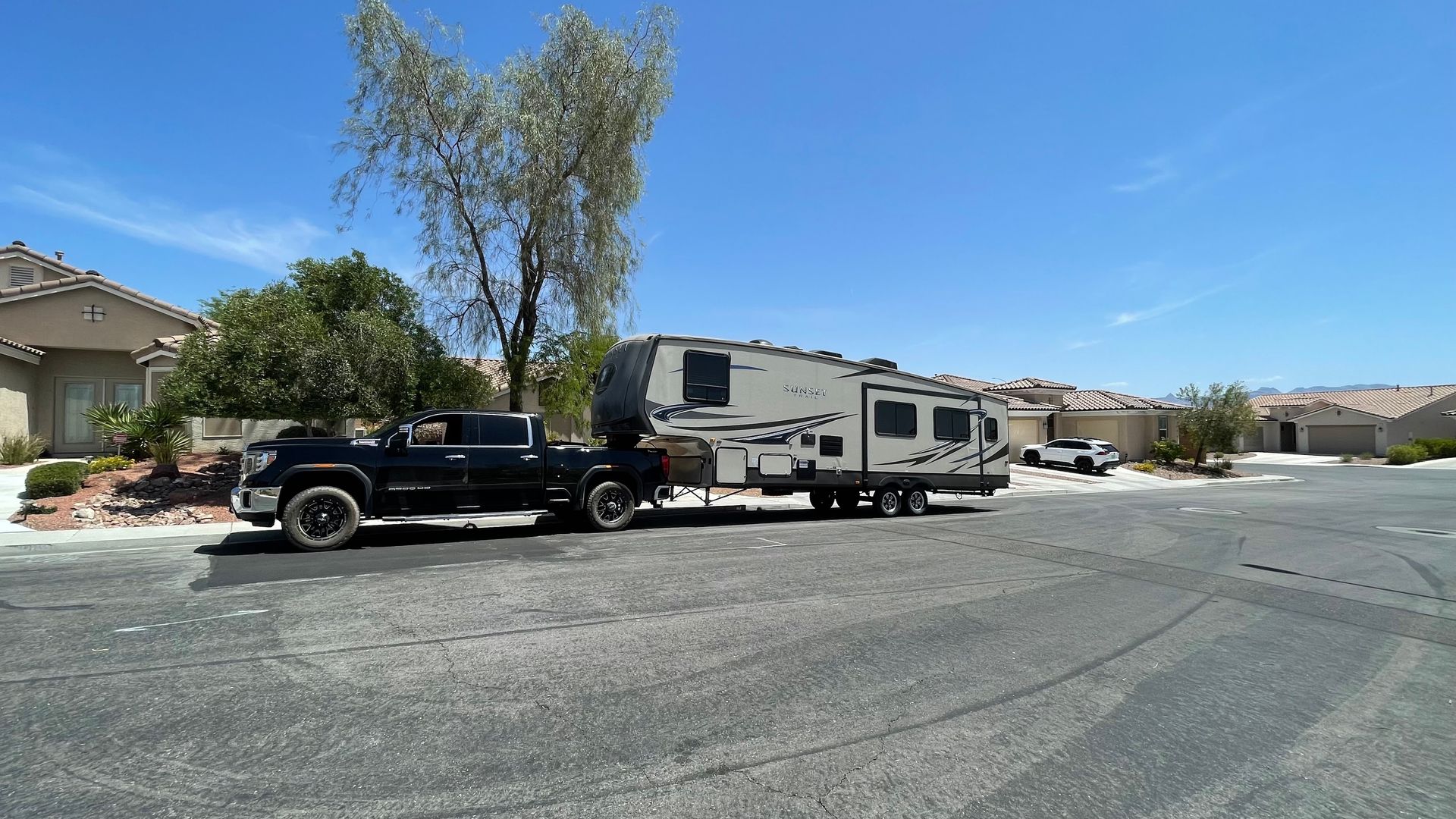 Black pickup truck towing a white RV on a suburban street under a clear blue sky