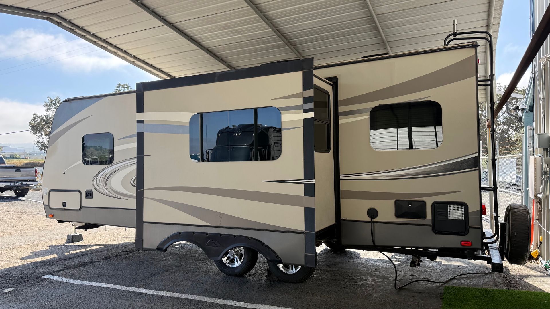 Beige and black RV parked under a carport, with an awning extended over the side.