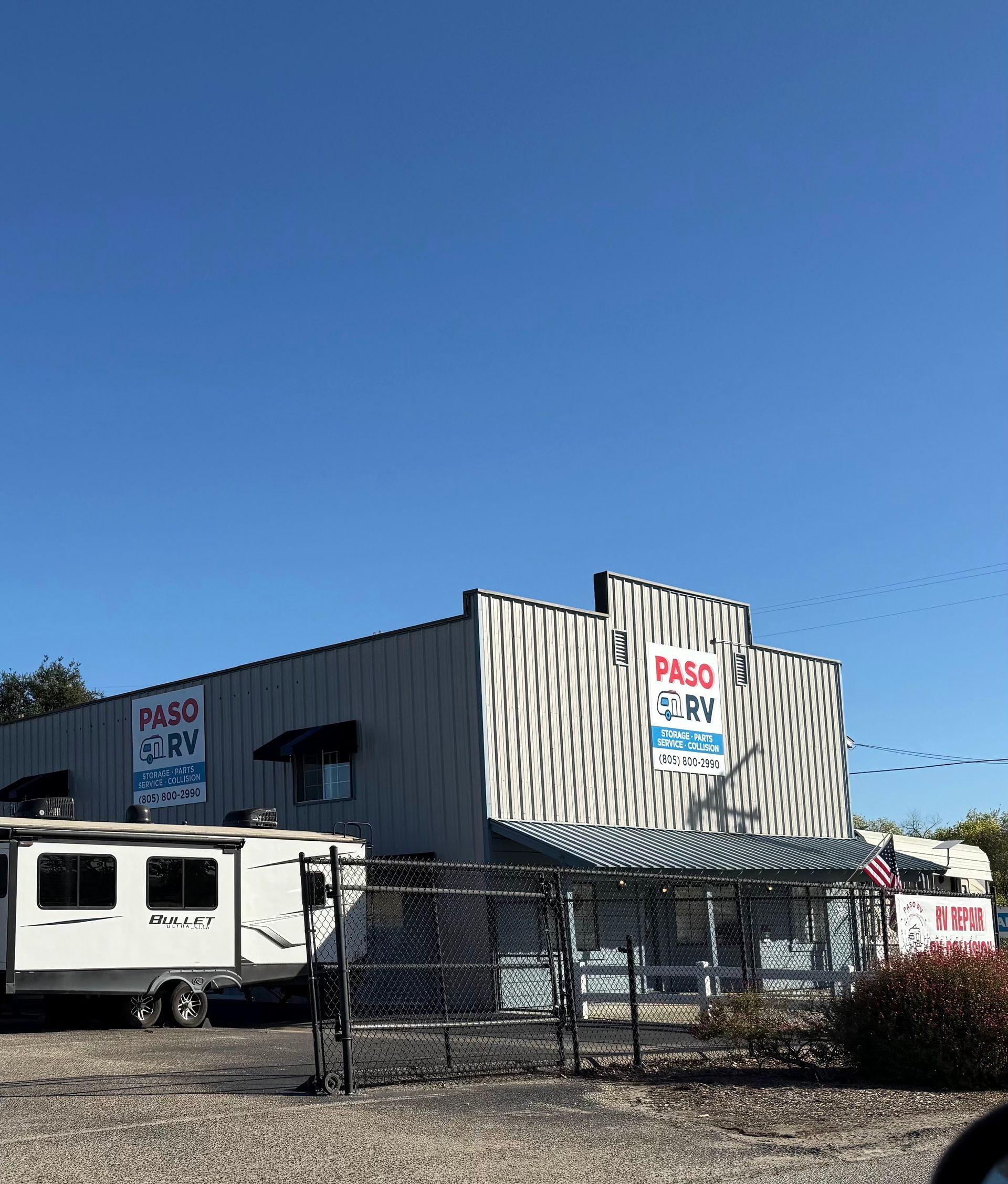 Commercial building with PASO signs, parked trailer, and chain-link fence under a clear blue sky